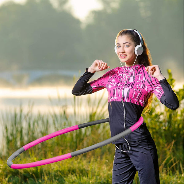 A young woman in a pink athletic outfit smiles while using the BRIT DIAMOND Exercise Hula Hoop for Adults—an adjustable, weighted fitness hoop for core and cardio workouts—outdoors near a lake surrounded by green vegetation.