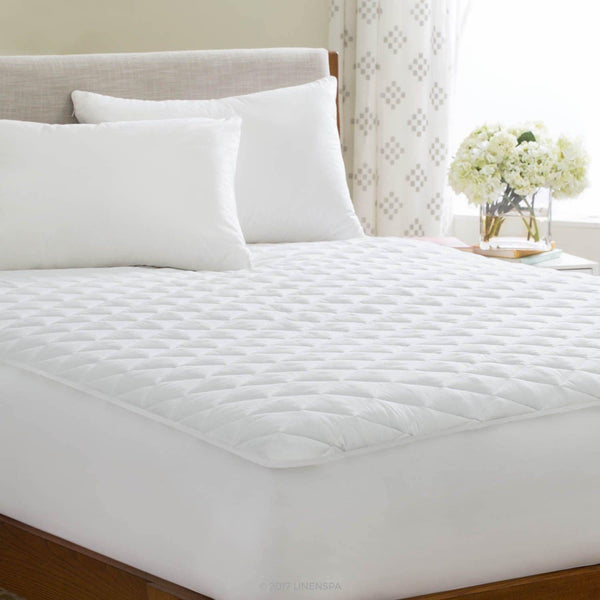 A neatly made bed features the BRIT DIAMOND Quilted Mattress Protector 100% Cotton (extra deep, all UK sizes), two large white pillows, and a smaller pillow beside a wooden nightstand with white flowers in a bright, airy room.