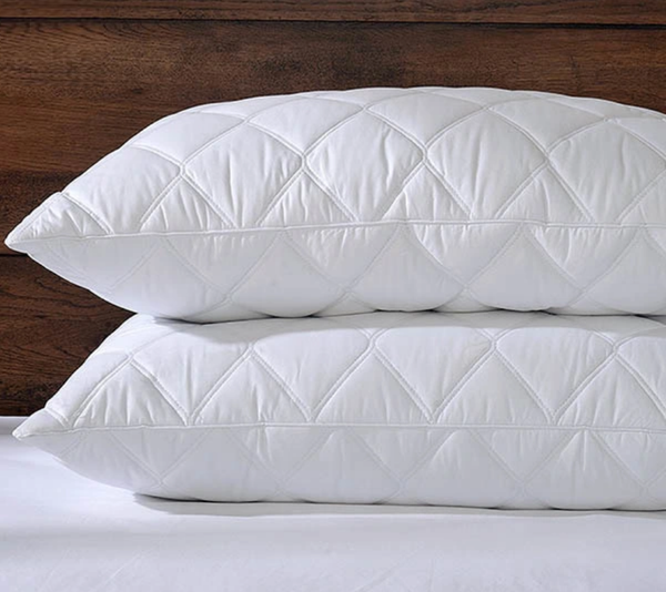 Two BRIT DIAMOND Luxury Quilted Hollow Fibre Pillows, extra soft, bounce back, and anti-allergic, are stacked on a bed with a wooden headboard in the background.