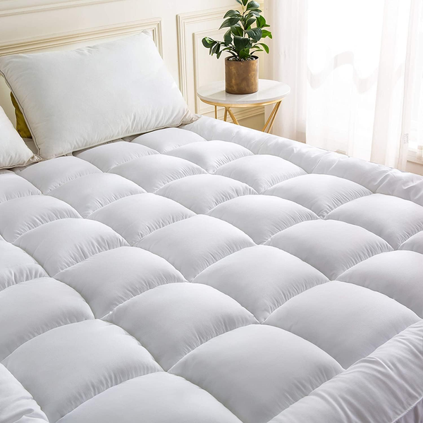 A neatly made bed features the BRIT DIAMOND Extra Deep Mattress Topper, 10cm thick and quilted, with two white pillows and a small potted plant on a round bedside table beside a window with sheer curtains.