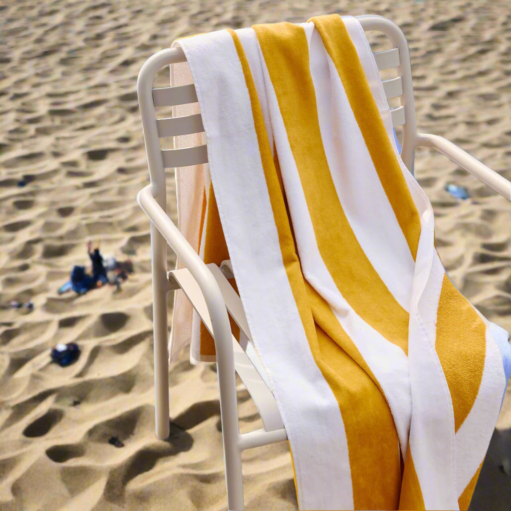 A BRIT DIAMOND - COMFORT IN EVERY STITCH Luxury Large Beach Towel, soft, absorbent, and quick-dry, in yellow and white stripes, is draped over a white metal chair on sandy beach with scattered debris. Pack of 2 available in multiple colours.