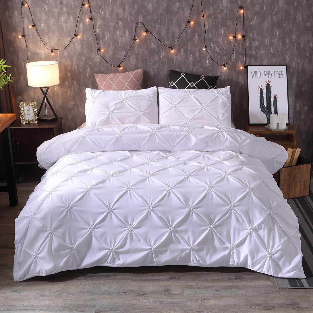 A cozy bedroom with the BRIT DIAMOND Pintuck Duvet Set in 100% Egyptian Cotton on the bed, pillows, string lights, a lit table lamp, and decorative items like a plant and framed art on the nightstand.