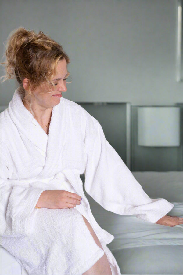 A woman in the BRIT DIAMOND Luxury Cotton Bathrobe – Ultra-Soft, Lightweight & Perfect for Everyday Use with an adjustable belt sits on a neatly made bed with white bedding, enjoying the breathable cottons comfort and relaxation.
