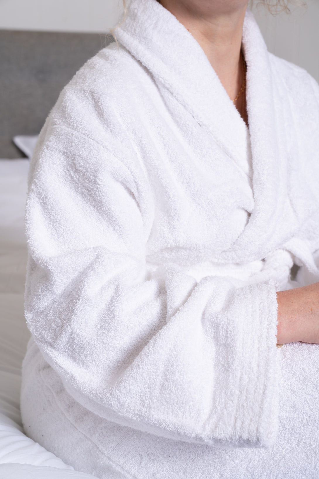 A person in a BRIT DIAMOND Luxury Cotton Bathrobe – Ultra-Soft, Lightweight & Perfect for Everyday Use sits on a bed, only their lower face and upper body visible. White bedding and the headboard accentuate the robe’s breathable cotton comfort.