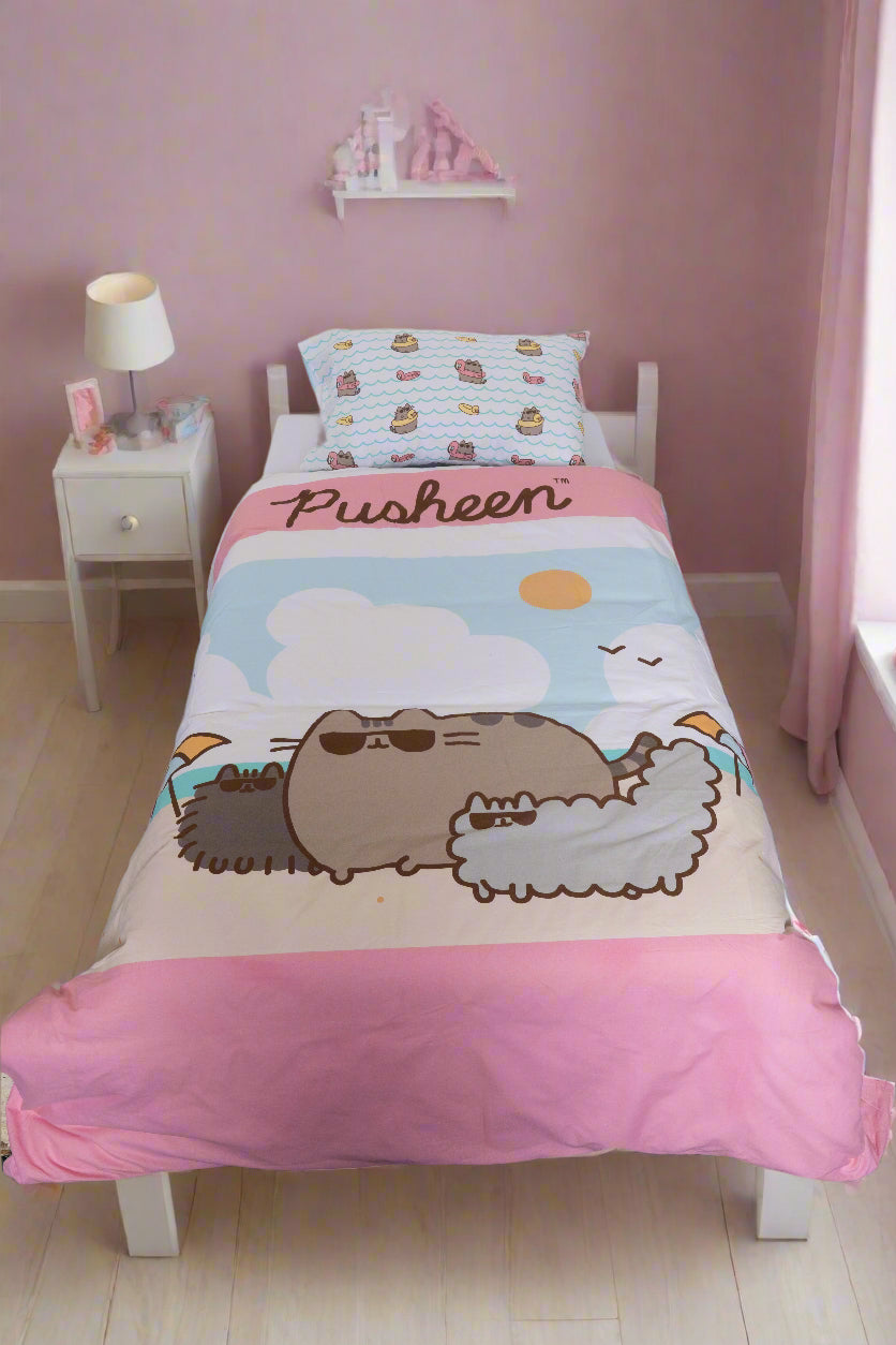 A cozy single bed with pink walls features the BRIT DIAMOND Kids Pusheen Reversible Duvet Cover Set—cute, double-sided bedding—next to a white nightstand with a lamp and decor in a sunlit bedroom.
