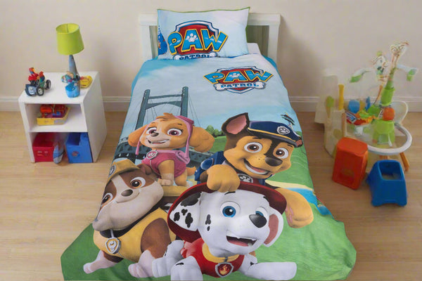 A children’s room features the BRIT DIAMOND Kids Paw Patrol Reversible Duvet Cover Set—2-in-1 Adventure Bedding for Little Heroes—with cartoon characters, a white nightstand, toys, lamp, and a playset with slide on a wooden floor.