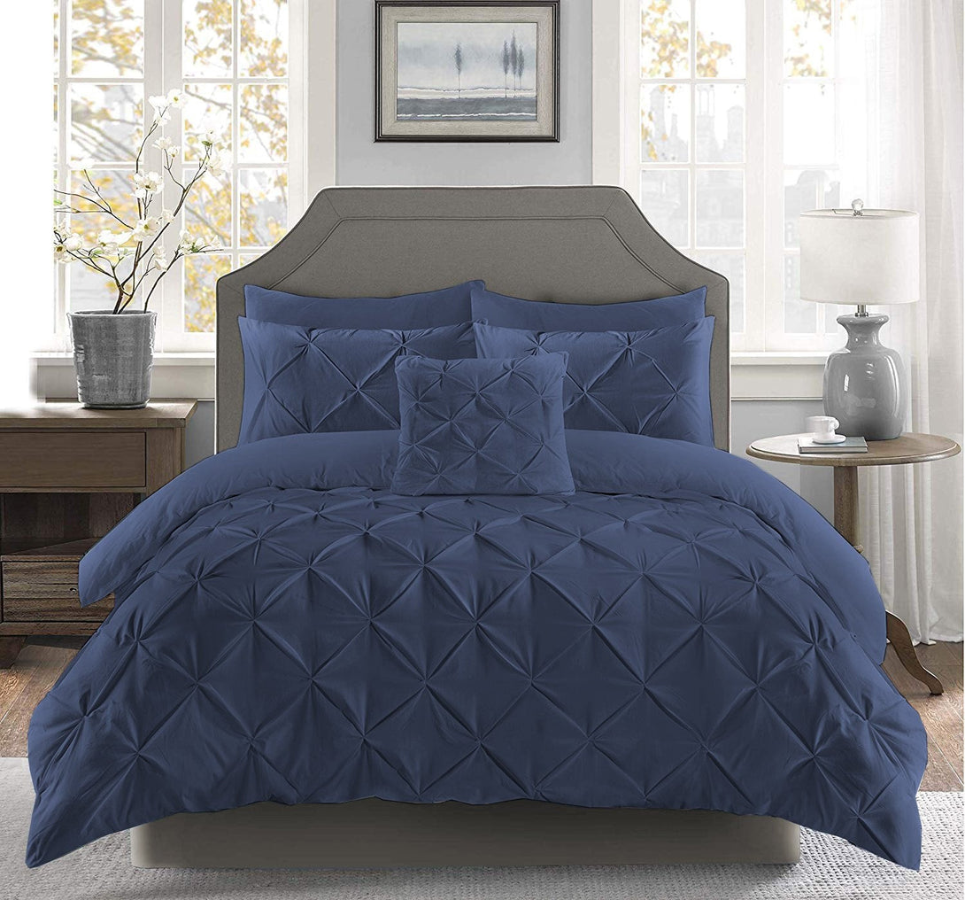 A bed features the BRIT DIAMOND Pintuck Duvet Set (100% Egyptian Cotton) in dark blue with matching pillows between two nightstands and lamps. A framed picture hangs above the headboard, and large windows are in the background.