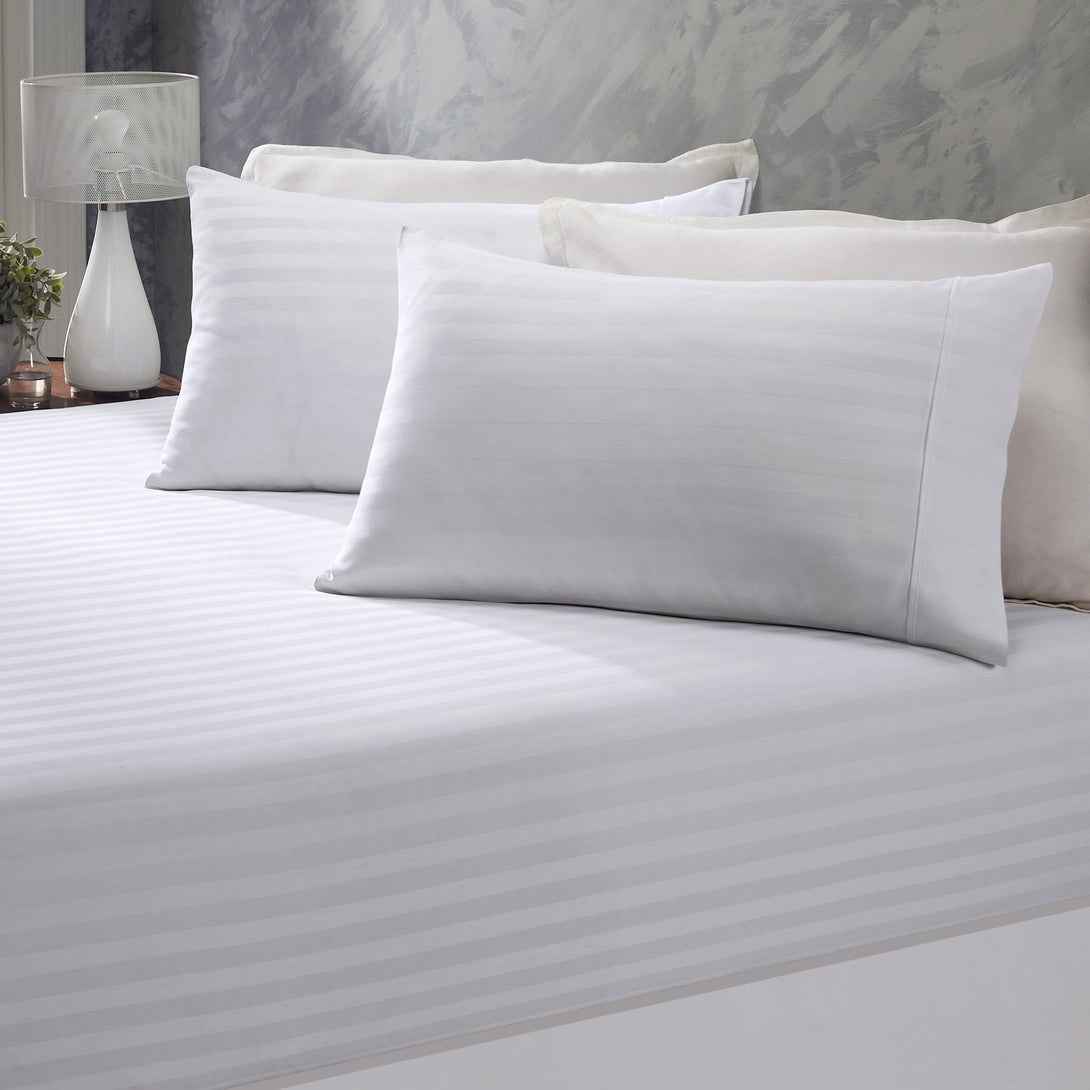 A neatly made bed features BRIT DIAMONDs Stripe Extra Deep Fitted Sheet (100% Egyptian Cotton, 400TC) with matching pillowcases, two large pillows, a modern lamp, and a small plant on a bedside table against a textured wall.