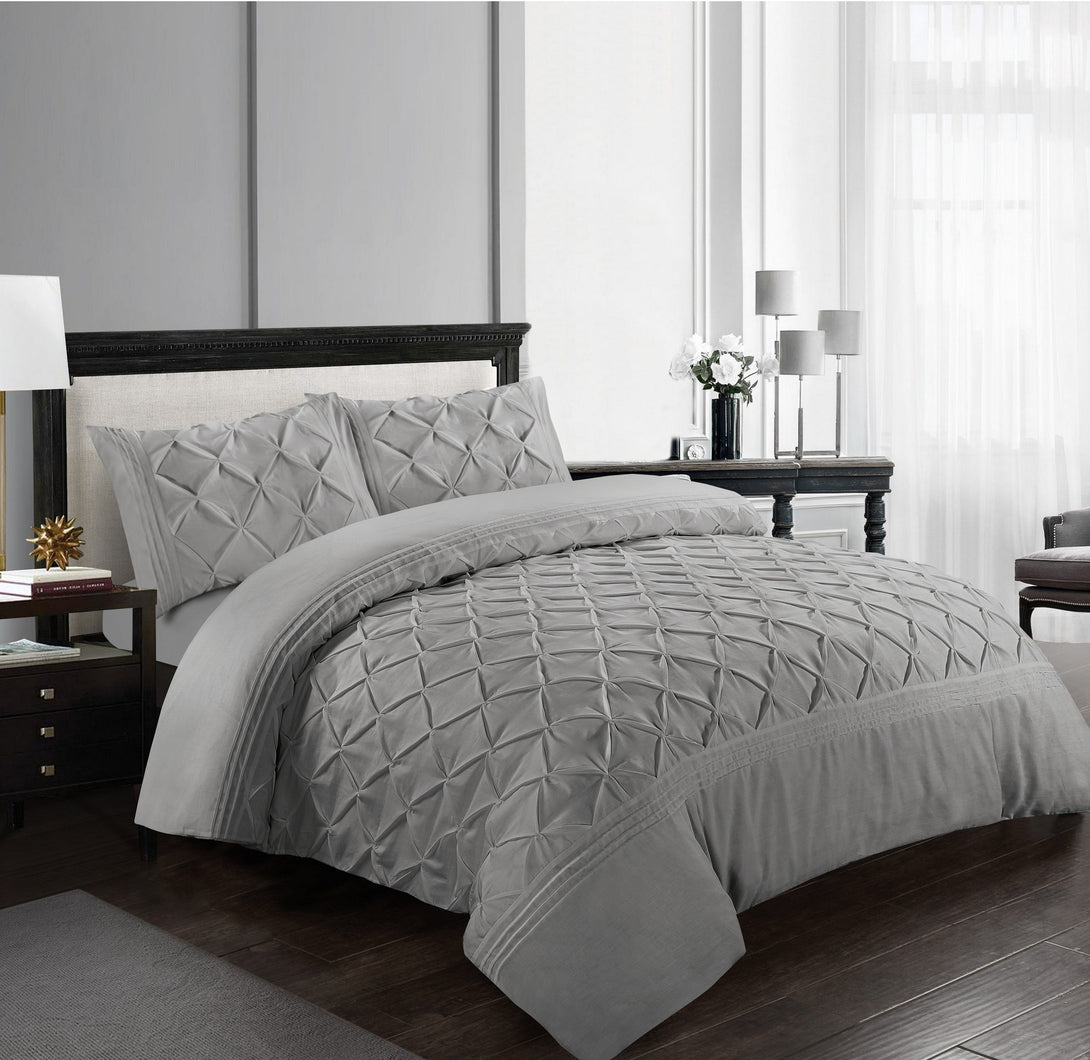 A modern bedroom features the BRIT DIAMOND Deluxe Pinch Pleated Duvet Cover Set, crafted from soft 100% cotton, with matching pillowcases. Dark wood furniture, a lamp, and a vase of white flowers accent the sunlit space with sheer curtains.