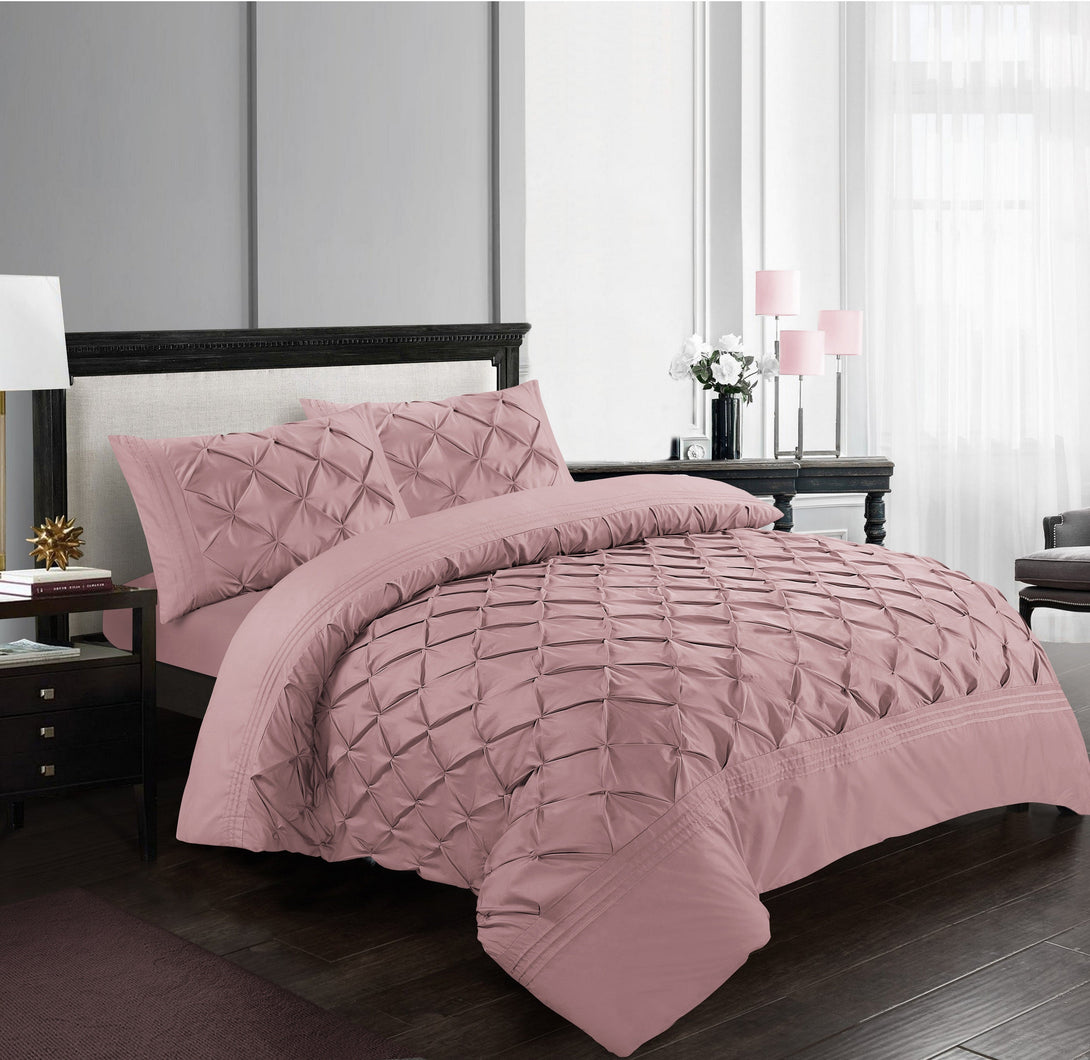 A neatly made bed features the BRIT DIAMOND Deluxe Pinch Pleated Duvet Cover Set in textured dusty pink 100% cotton, with matching pillowcases. The room has dark furniture, a lamp, and large windows with sheer white curtains.