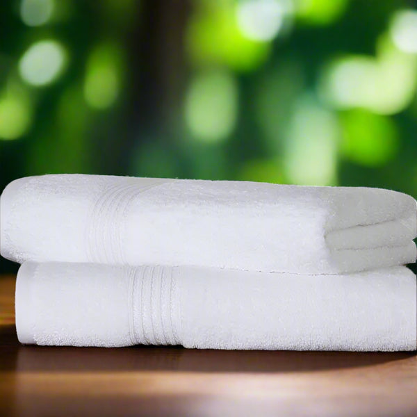 Two BRIT DIAMOND 100% cotton bath sheets, quick-dry and ultra soft, are neatly stacked on wood, with green plants and a white brick wall in the background. Ideal for spa, gym, guest, or bathroom. Pack of 2 premium towels.
