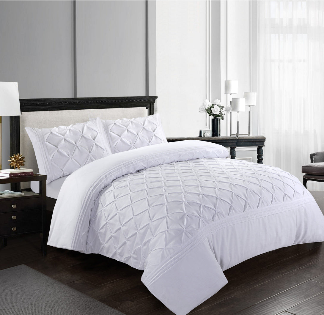 A modern bedroom features the BRIT DIAMOND Deluxe Pinch Pleated Duvet Cover Set—soft 100% cotton pintuck bedding with matching pillowcases—paired with a black headboard, dark wood furniture, tall windows, and white decor accents.