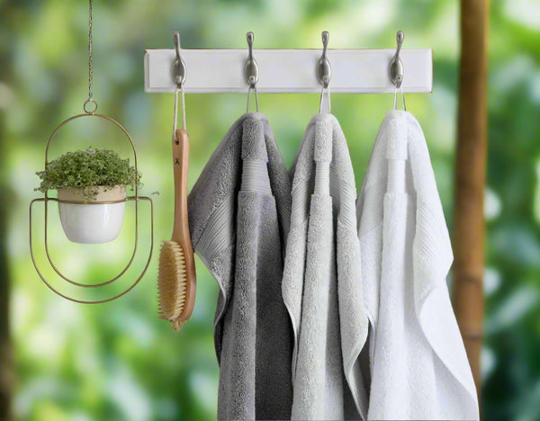 A hanging rack with four metal hooks holds a wooden bath brush and three BRIT DIAMOND 100% cotton quick-dry bath towels in white and gray. A small potted plant hangs nearby in a round gold holder, set against a blurred green background.