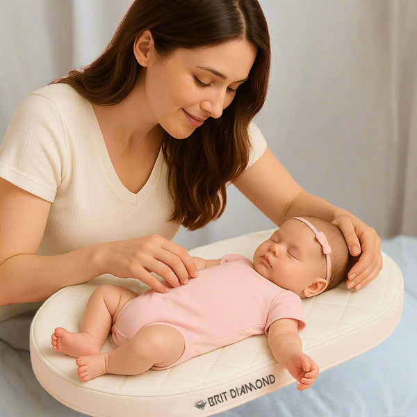 A woman gently touches a sleeping baby in pink, resting on the BRIT DIAMOND Premium Baby Moses Basket Foam Mattress—soft, breathable, and safe for newborns—atop a white cushioned pad against a soft pink background.