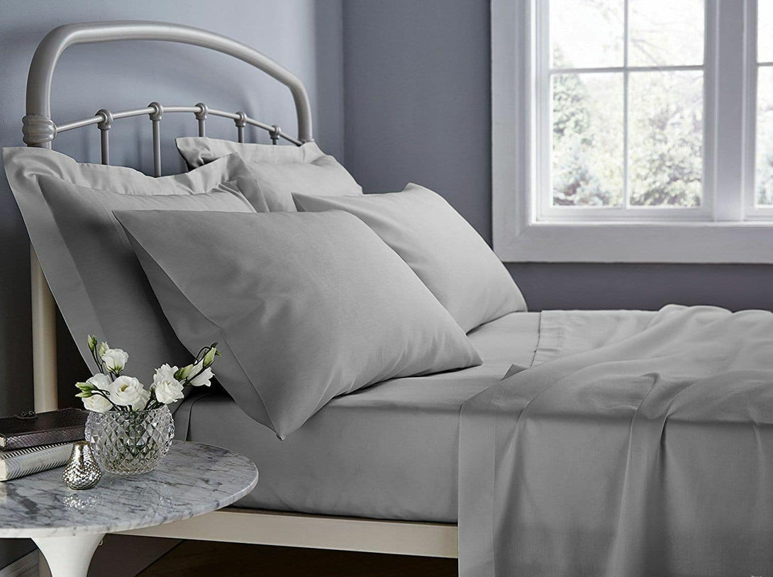 A neatly made bed with BRIT DIAMOND Luxury Hotel Quality 400TC Extra Deep Fitted Sheet in soft gray sits by a window. A marble nightstand holds a vase of white flowers and a decorative bowl, complementing the rooms serene gray walls.