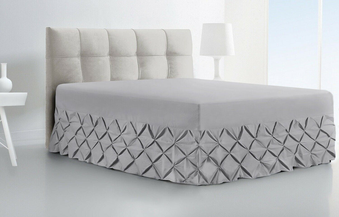A modern bedroom featuring a BRIT DIAMOND Luxury Fitted Pintuck Valance Sheet in 100% Egyptian cotton, with a light upholstered headboard, next to a white nightstand and tall white lamp.