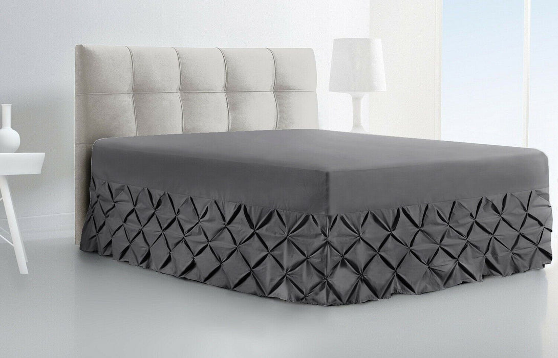 A modern bedroom features a beige tufted headboard, BRIT DIAMOND Luxury Fitted Pintuck Valance Sheet (100% Egyptian Cotton) in dark gray, a white nightstand, and a white lamp in a minimalist setting.