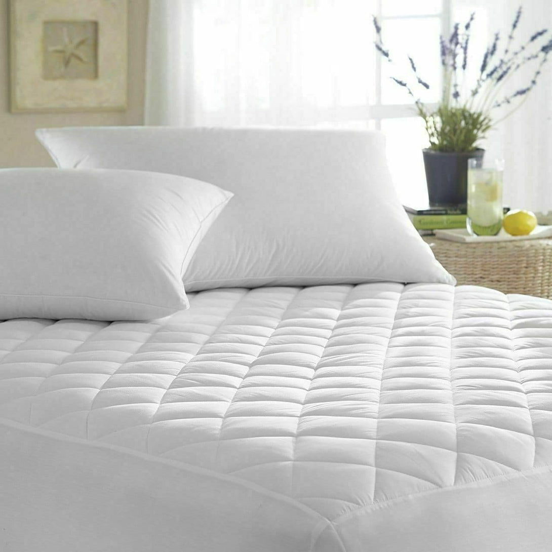 A neatly made bed showcases the BRIT DIAMOND Quilted Mattress Protector 100% Cotton, a fitted bed cover available in all UK sizes with 30cm & 40cm extra deep options. In the background, sunlight streams by a wicker table with lemonade and lavender.