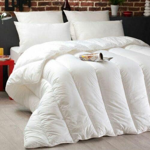 A neatly made bed features the BRIT DIAMOND Premium Hotel Quality Duvet Deep Sleep Quilt, surrounded by white pillows. A red nightstand stands beside the bed against a brick wall, with a magazine resting on the quilted comforter.