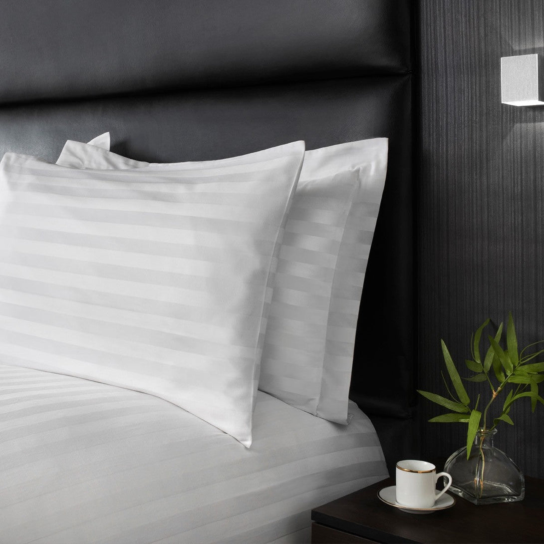 A neatly made bed with two white striped pillows and a BRIT DIAMOND Luxury 400 Thread Count Satin Stripe Duvet Set in 100% Egyptian Cotton, next to a dark nightstand with a small plant in a glass vase, coffee cup, and black padded headboard.