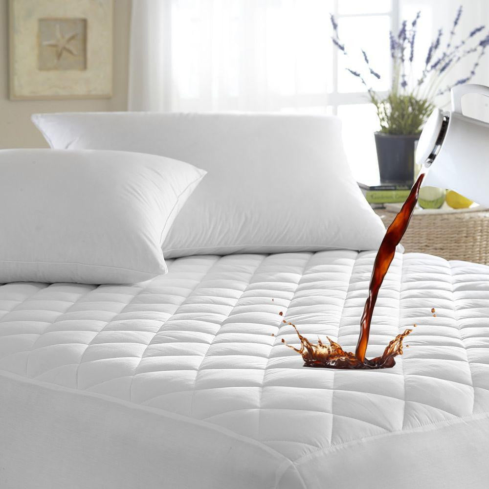 A stream of dark liquid is being poured from a white pot onto the BRIT DIAMOND Extra Deep 100% Waterproof Quilted Mattress Protector on a neatly made bed with white pillows in a bright, airy bedroom.